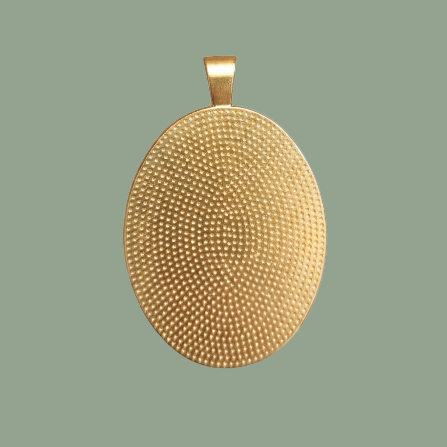 A gold-toned solid brass oval pendant 'blank' with a detailed dotted textured surface, presented against a seamless sage green background, showing the bail at the top.